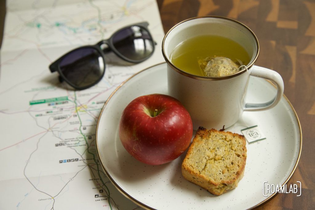 A slice of lemon poppyseed squash bread on a plate with a red apple and mug of tea set on top of a map.