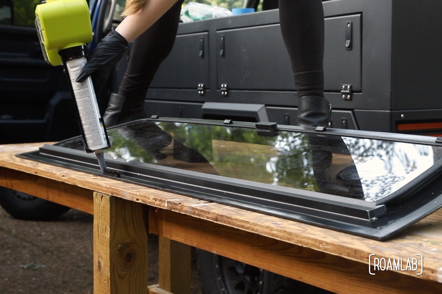 Woman standing on a wood work table using an application gun to lay a bead of urethane on a trucks rear windshield.