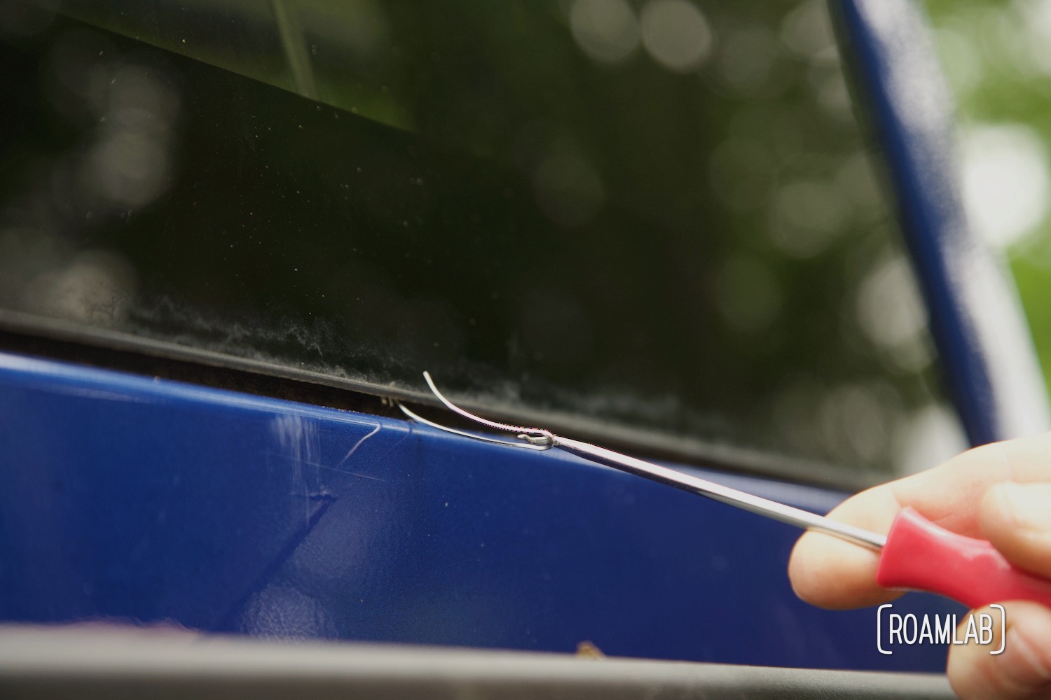 Hand pulling a hook and cutting wire through a gap between the black windshield glass and blue metal frame.