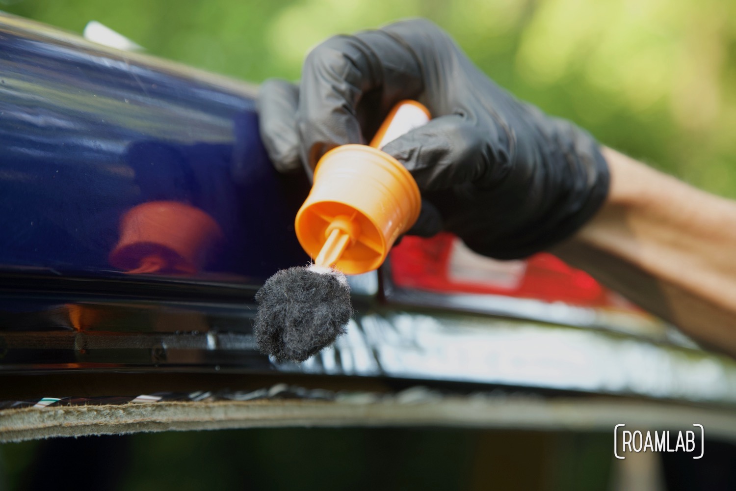 Gloved hand using an orange dauber to apply black automative glass primer on the blue truck frame.
