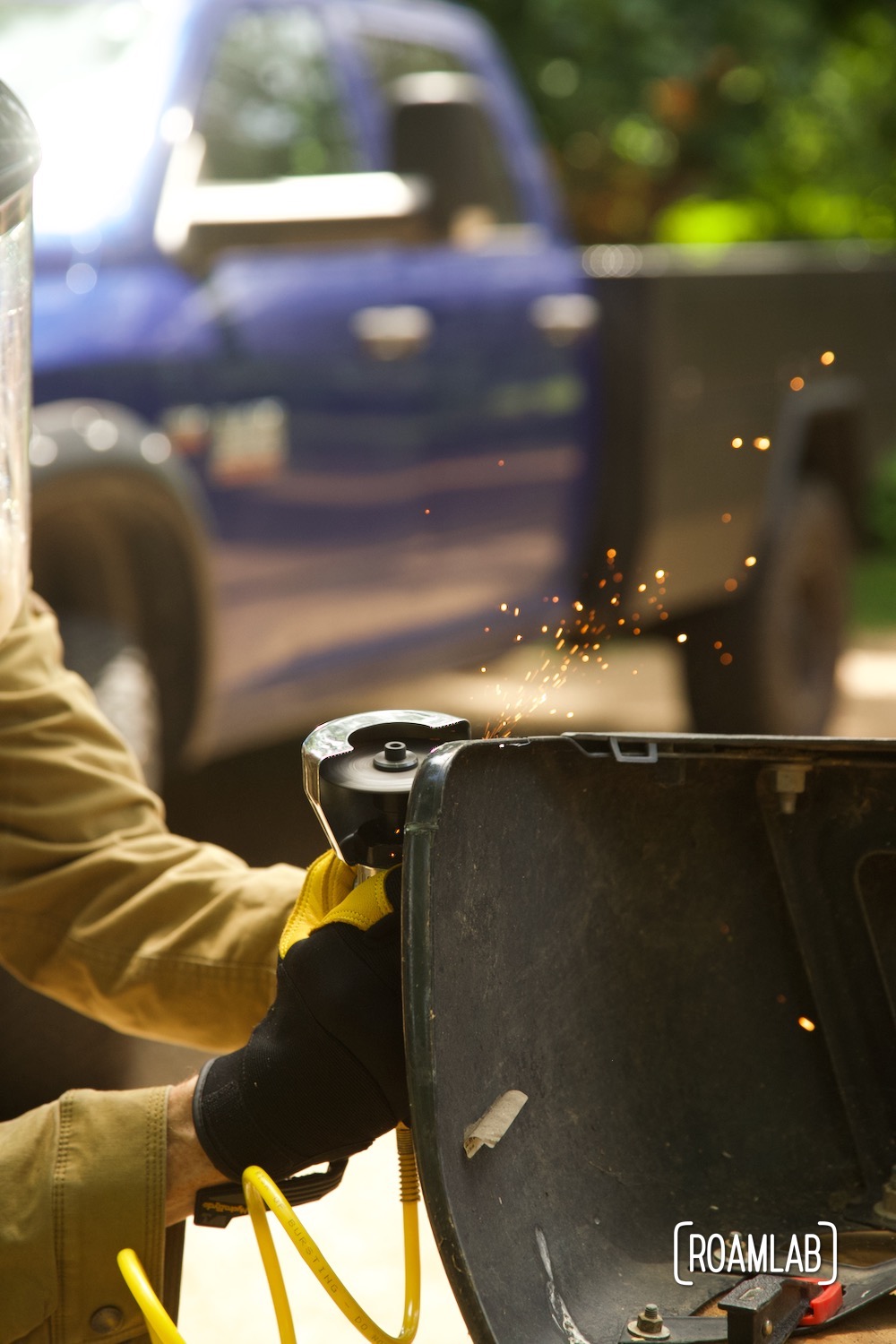 Gloved hands using a cut-off tool to trim a truck bumper with a truck parked in the background