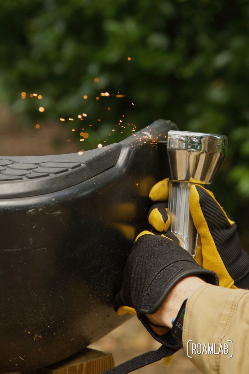 Close up of gloved hands using a cut-off tool to trim a truck bumper with bushes in the background.