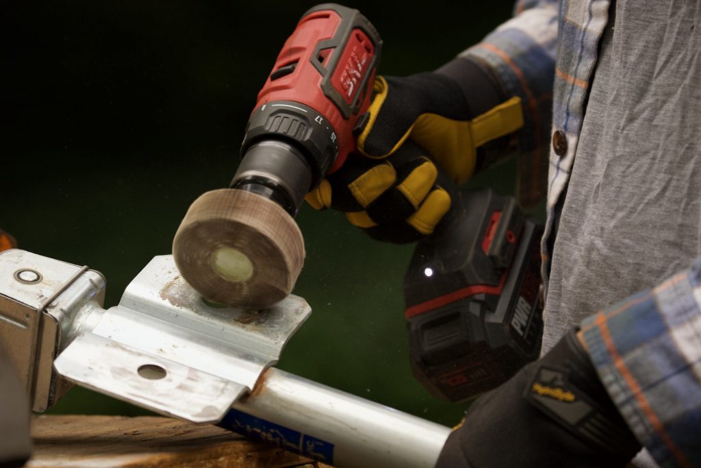 Gloved hands holding a power drill with an abrasive flapper wheel attachment to grind off corrosion on the mount point of a steel jack.