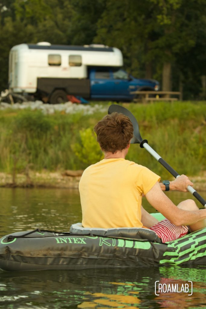 Man kayaking towards a truck camper parked on the shore.