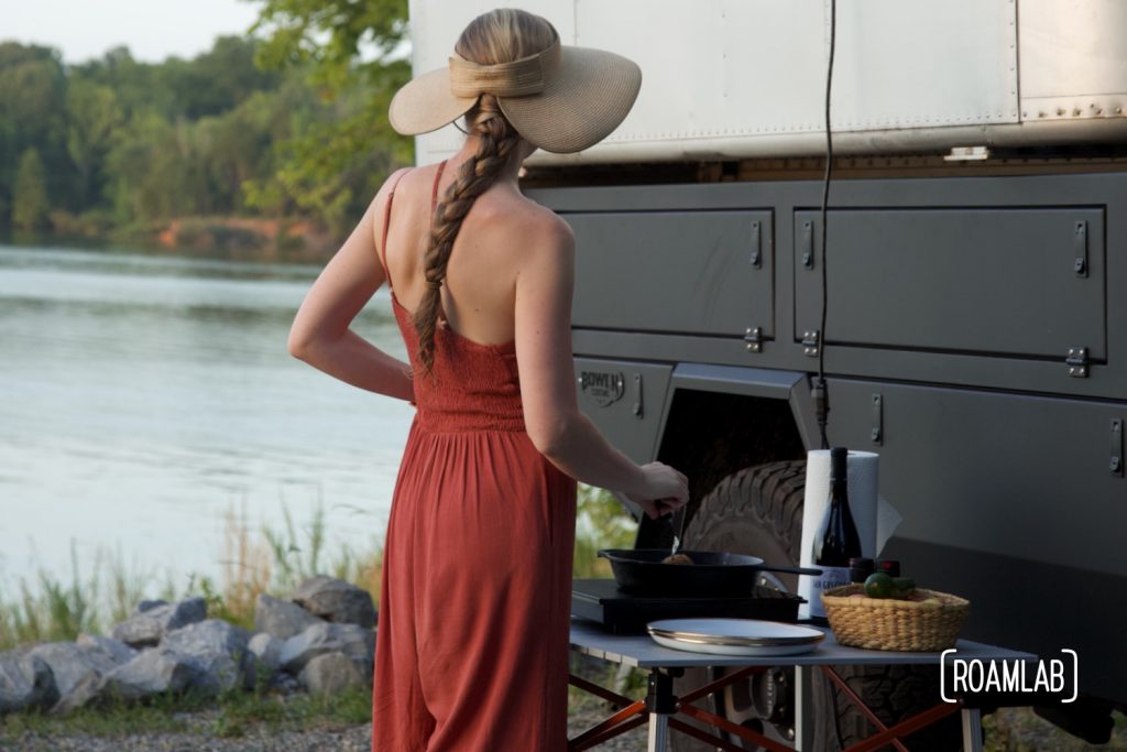 Woman standing next to a truck camper, cooking on a skillet, next to a body of water.