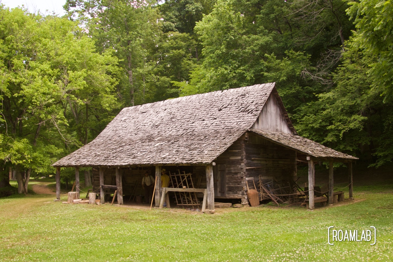 Large wood shingle log tool barn sitting in a grass field with surrounded by woods.