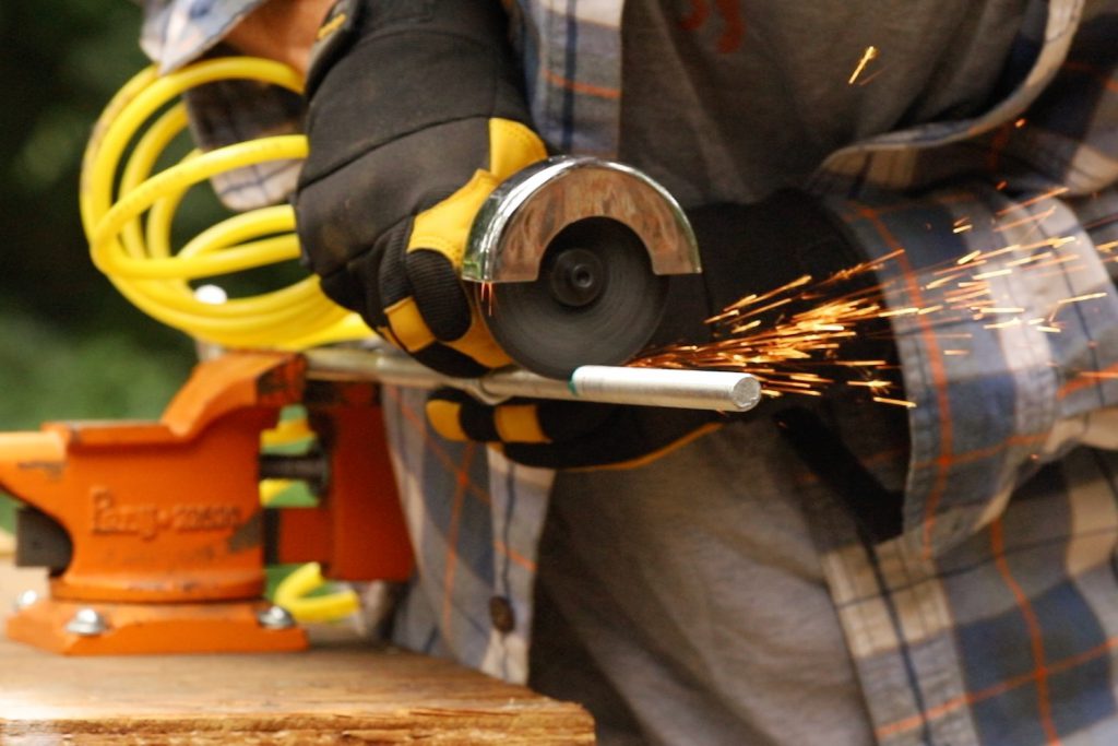 Threaded metal rod secured in an orange worktable vice as a man sends sparks flying with a cut-off tool.