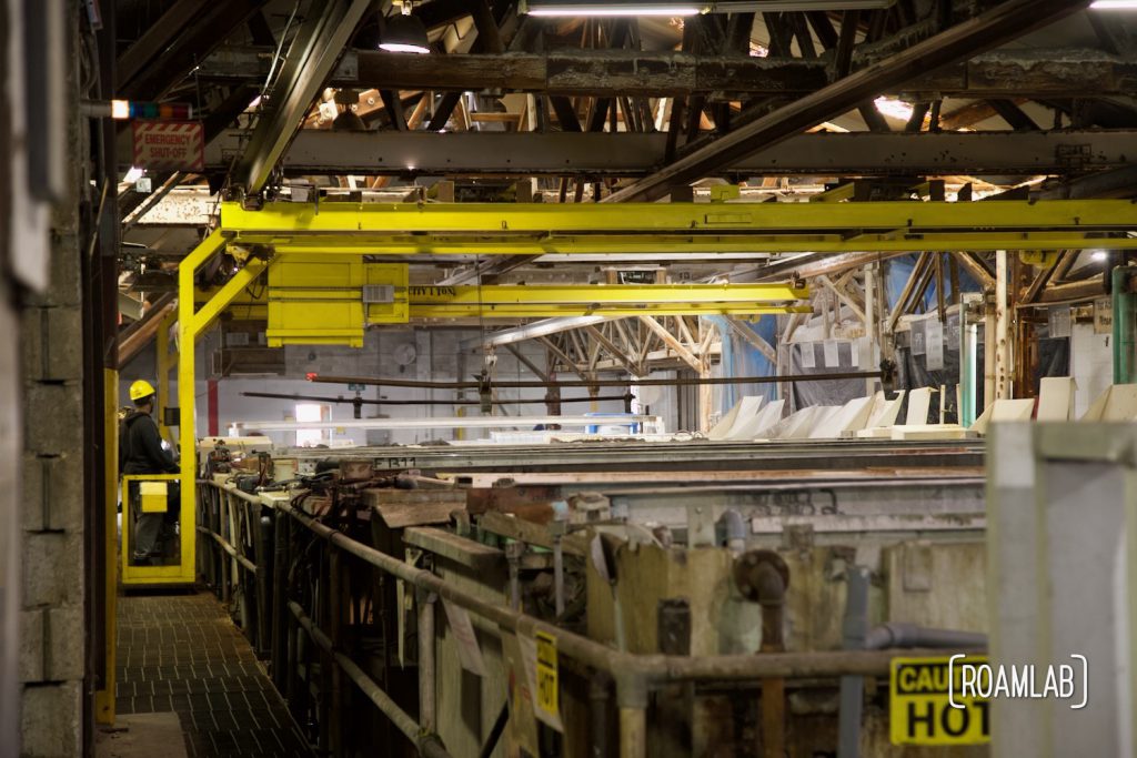 View inside a large warehouse filled by a row of vats and a yellow crane straddling  the row.