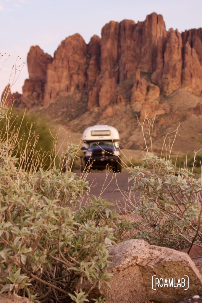 Sage plants hiding a 1970 Avion C11 truck camper in front of the Superstition Mountains in Lost Dutchman State Park Campground at sunset.