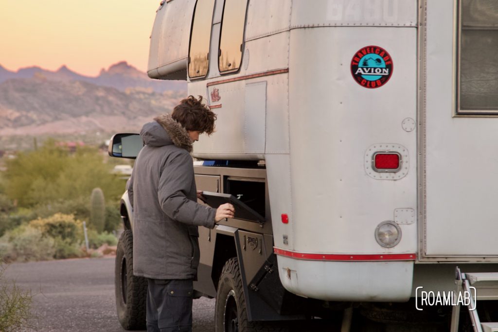 Man opening a truck bed box on a 1970 Avion C11 truck camper at sunrise.