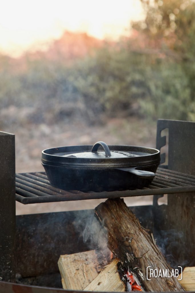 Covered cast iron skillet over a campfire with a red desert mountain in the background.