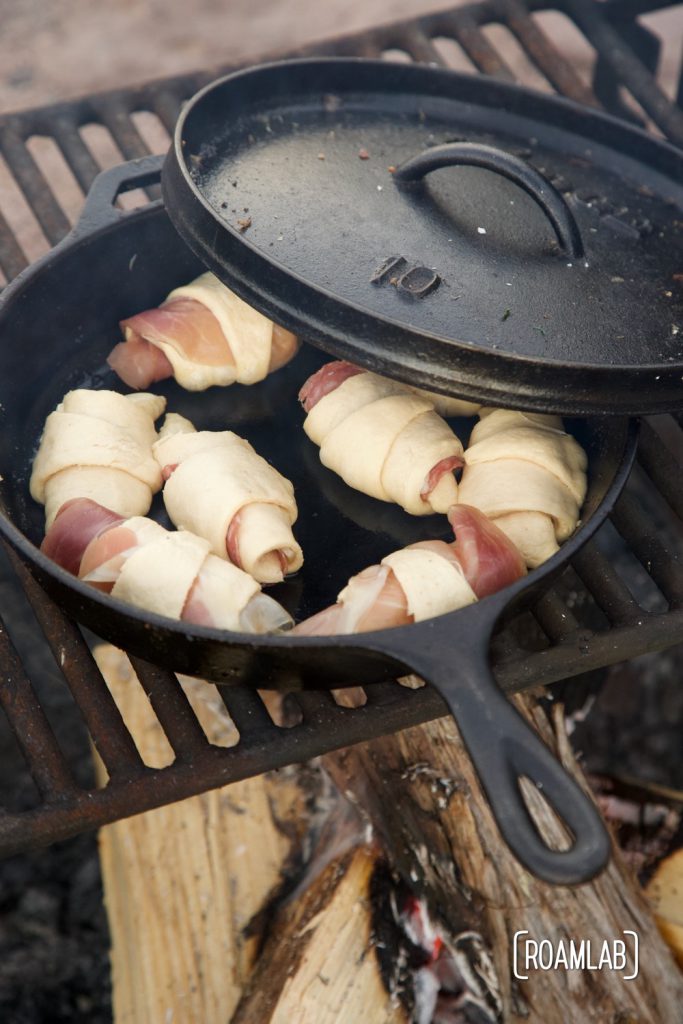 Prosecute  and cheese mini croissants baking over a campfire in a cast iron skillet.