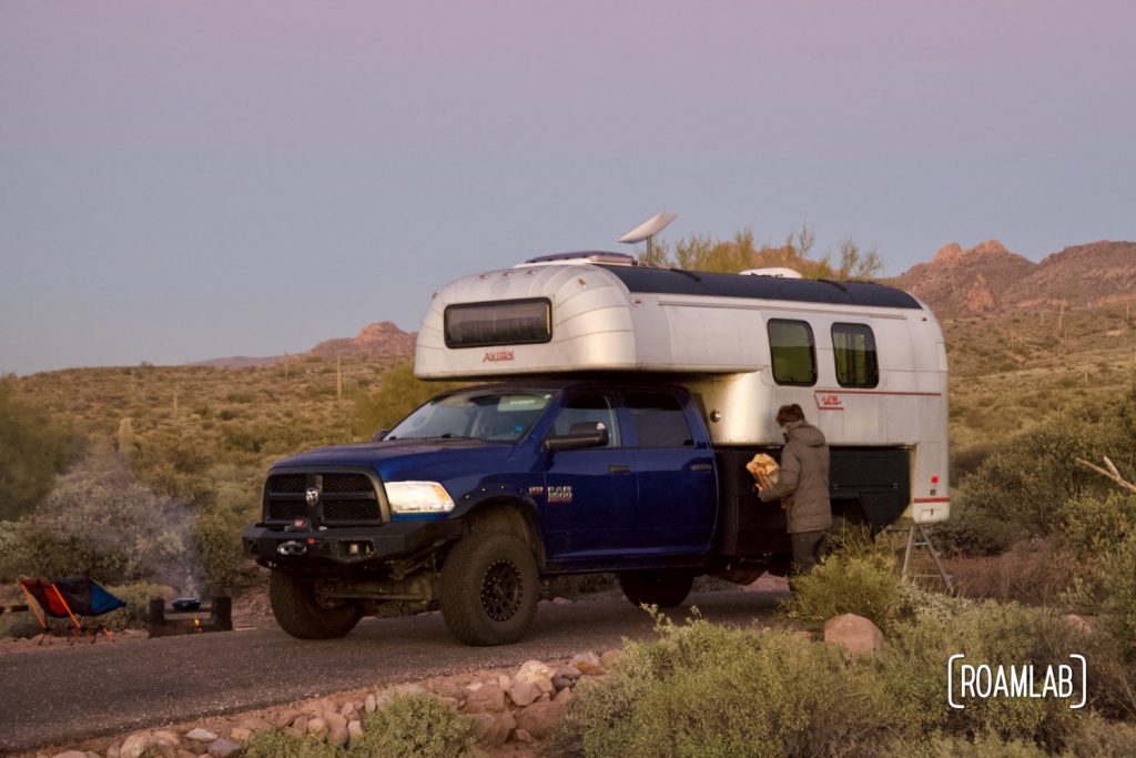 Man pulling a bundle of wood out of a truck bed storage box, part of a vintage 1970 Avion C11 truck camper parked in the desert at sunset.